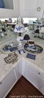 Photo showing full setup of blue and white porcelain items including table lamps, serving trays, plates, utensils, and metal trivets on white marble counter with mirrored background.