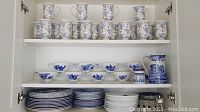 Three-shelf cabinet overview showing top shelf footed mugs, middle shelf handled bowls, pitcher and jar, bottom shelf stacks of plates