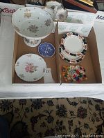 Full view of all items in lot displayed in box. Shows pedestal fruit bowl, white floral plate, blue decorative plate, set of three blue and orange floral border plates, and assorted glass candies in small box.