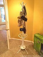 Full view of white metal coat rack with six handbags hanging, against yellow wall and carpeted floor.
