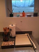 Photo of a countertop with wooden utensils holder, paper towel holder with white paper towel, rotary paper cutter, and a decorative box.