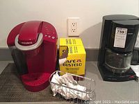 Photo showing red Keurig coffee maker, box of Cafe Bustelo coffee pods, small wire basket with coffee pod sleeves, and black Krups drip coffee maker with glass carafe.