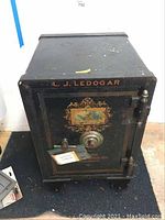 Front view of the black metal safe showing the decorative gold and red design with an image on the door, and the company name L. J. Ledogar printed above the door.