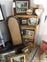 Several framed artwork pieces stacked against a wall and a wooden shelf with five tiers holding multiple framed pieces including landscapes and a heart-shaped frame.