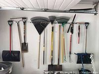 Wide view of gardening tools hanging on wall including shovels, rakes, hoes, and hedge shears