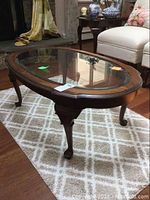Oblong glass top coffee table placed on light-colored patterned rug with wooden cabriole legs and beveled glass inset top.