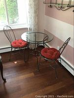 Front angle view of the glass top bistro table with two wrought iron chairs with red-orange cushions near a window and striped wall
