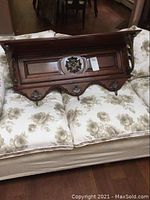 Wooden brown shelf with decorative carved panels and bronze tone metal hooks attached beneath, shown lying on floral patterned cushions.