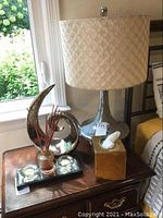 Photo of a 24-inch chrome metal lamp with patterned fabric shade, abstract chrome metal swirl sculpture, black mirrored tray with three glass votive candles, and a decorative jar with reed diffuser sticks on a wooden table.