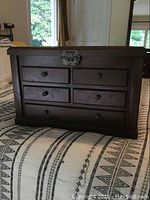 Front view of a dark wooden jewelry box with five drawers and a metal lock on top, placed on a bed with patterned bedding.