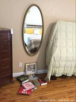 Oval wall mirror hanging on wall beside bed and dresser, with books and references placed on floor below it.