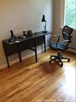 Side view of black drop leaf computer desk with items on top (not included) and swivel office chair beside it on wooden floor against white wall.