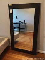 Full view of the rectangular wall mirror standing on the floor against a wall, showing the dark textured frame and clear reflective surface.