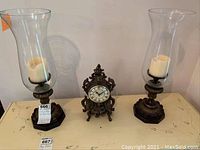Two metal candle holders with clear glass chimneys and white pillar candles, next to an ornate bronze-colored metal clock with Roman numerals on its face, all placed on a light-colored surface.