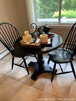 Wide view of black round table with decorative pedestal base and two Windsor chairs around it, with candles and other small items on table
