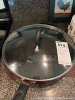 Black nonstick frying pan with glass lid and black handle, sitting on a granite countertop next to a framed rooster picture.