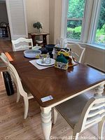 Wooden farmhouse dining table with rectangular brown top and white painted legs. Four ladder back chairs surround the table, beige cushioned seats visible. Various items on table excluded from sale.