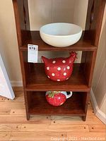 Three ceramic home decor items displayed on wooden shelving: red polka dot chicken figurine, white bowl, strawberry pitcher.