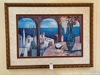 Framed wall picture featuring a coastal scene with arches, buildings, ocean, and a vase with flowers inside a gold-tone detailed frame.