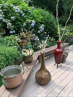 Photo showing two standing plant holders with metal pots on a deck, a large decorative standing vase with artificial white flowers, and a tall red standing vase with artificial twisted branches, surrounded by garden foliage.