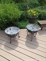 Two round metal planters on decorative metal stands on a wooden deck with greenery in the background