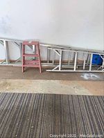 Photo showing three ladders side by side against a white wall on brown flooring: one red 2 ft step ladder, one medium 5 ft aluminum ladder, and one long 8 ft aluminum ladder.