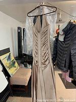 Beige satin halter dress hanging on hanger with transparent garment protector. Chair and artwork seen in background.