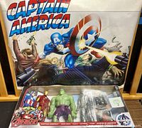 Photo of five 12-inch Marvel Avengers Titan Hero action figures in original blister packaging with Captain America poster in background, showing front view of figures and packaging details.
