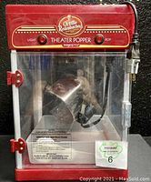 Front view of Orville Redenbacher Presto Theater Popper popcorn machine with plugged-in power cord, red control switches labeled Light and Pop, clear plastic door with red hinges and latch.