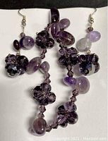 Close view of amethyst stretch bracelet and matching pierced earrings set lying on a black surface, showing various shapes of amethyst and faceted purple crystal flower beads.
