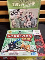 Golden Triviagame M.A.S.H Edition 1984 board game box and Monopoly Junior 2013 box shown from top, boxes stacked