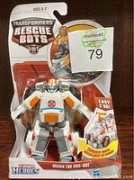 Two images each showing a brand new Transformers Rescue Bots Medix The Doc-Bot figure in sealed packaging. Figures are white and gray with orange details and are designed to convert from ambulance to robot as stated on packaging.