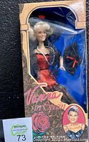 Full packaged doll with visible tear in clear plastic film of packaging, shows red satin dress and black lace overlay inside box with rose graphic.