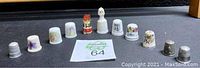 Front view of all 11 assorted vintage collectible thimbles arranged in a row on a black surface, showing various sizes, designs, and colors including porcelain, bone china, and metal.