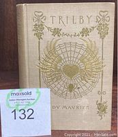 Front cover of the 1895 Trilby book showing gold embossed spider web design and title with seller lot number tag in front.