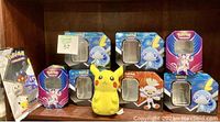 Eight Pokémon collectible tins arranged with visible artwork including Sobble, Scorbunny, Zacian, and a small Pikachu plush toy positioned in front.