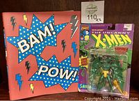 Photo showing Marvel X-Men Sauron action figure in original packaging next to trading cards binder with comic text 'BAM! POW!' and lightning bolts.