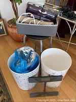 Gray caddy holding Conair Infiniti Pro curling irons, black stool, and two waste bins (white with pattern and white marble) with a blue plastic bag.
