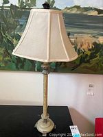 Full view of the metal table lamp with fabric shade placed on black surface in front of a landscape painting.