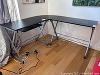 Full view of L-shaped black composite top corner desk with silver metal frame and cable management hole in corner, placed in room with hardwood floor.