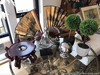 Wide shot showing entire set on glass tabletop including two Asian fans, two faux plants, covered candy dish on stand, seated metal figure, silver spherical object, and metal easel