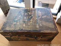 Top view of antique Asian trunk showing painted scene of figures outside, with visible wear and chipping on edges.