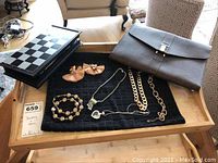 Full view of all items including five necklaces laid on black towel, tassel earrings, travel chess set box, and brown portfolio case on wooden tray.
