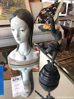 Porcelain bust and metal acrobat figurine displayed on glass table, with price tag visible on bust.