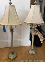 Pair of matching tall metal base table lamps with cream-colored fabric shades featuring bead fringe detail, shown side by side on wooden floor.