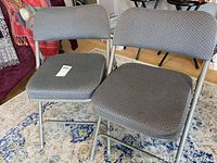 Front view of two gray patterned upholstered folding chairs sitting side by side on rug, metal frame visible.