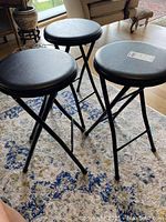 Three black folding stools on a patterned rug with cushioned seats and metal legs.