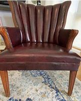 Front view of the burgundy leather-like armchair showing the fluted backrest, wooden legs, armrests with nailhead trim, and seat upholstery.
