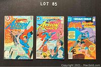 Photo showing 3 comic books on a dark surface marked Lot 85. The comics are Action Comics (2 issues) and Brave and the Bold (1 issue) featuring DC characters Superman, Batman, and Wonder Woman. All comics show visible wear and are unbagged.