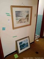 Two framed landscape paintings positioned near a wall; one hanging and one leaning on the floor
